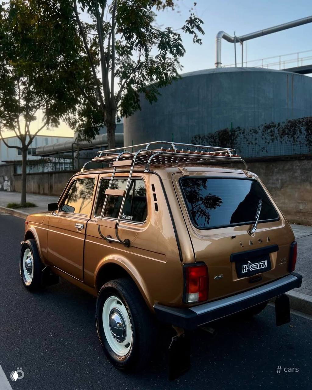 Задний ракурс золотистой Нивы на городской улице: экспедиционный багажник на крыше из дерева и металла, классические хромированные детали.