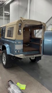 Задняя часть ретро-стилизованного кемпера на базе внедорожника: открытый багажник с деревянным полом, брезентовая крыша и большие внедорожные колёса в ангаре.