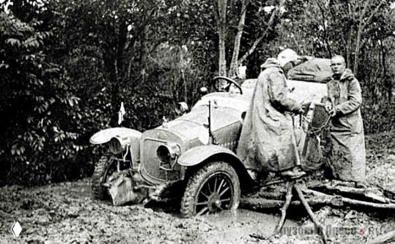 Чёрно-белая фотография раннего автомобиля, застрявшего в грязи; рядом мужчины в прежней одежде помогают вытянуть машину.