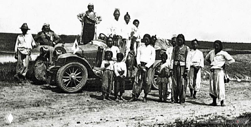 Группа людей и ретро-автомобиль на равнине; вокруг местные жители в традиционной одежде наблюдают за экипажем.