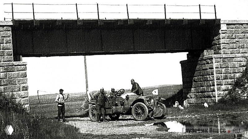 Ретро-автомобиль остановлен под железнодорожным мостом, рядом несколько человек обсуждают дальнейший маршрут.