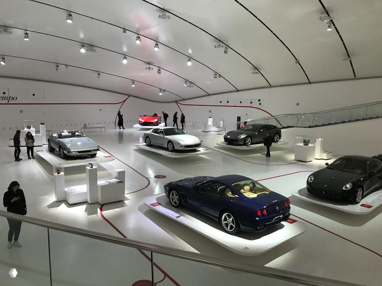 Большой зал музея с экспонатами Ferrari: выставочные подиумы с классическими моделями и просторный белый интерьер с посетителями.