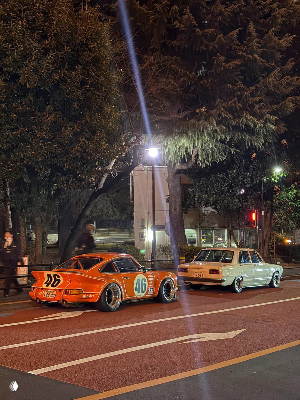 Две ретро‑машины — оранжевый гоночный Porsche‑стайл с номером 46 и белый старый седан, припаркованные ночью вдоль улицы на фоне деревьев и фонарей.