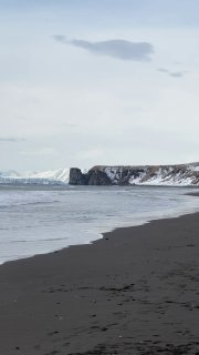 Пустынный берег Камчатки с чёрным вулканическим песком, скалистыми утёсами и серым морем под облачным небом.