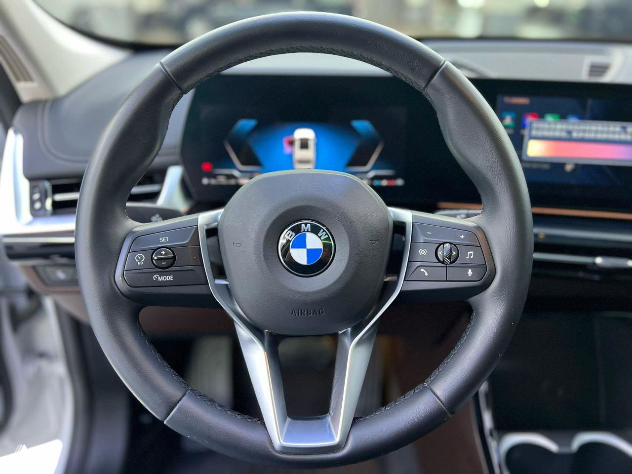 Крупный план рулевого колеса BMW с эмблемой и кнопками управления, на заднем плане — цифровая панель и информационный дисплей салона.