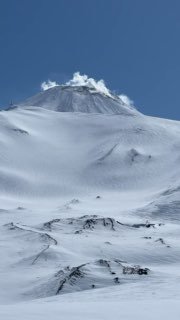 Заснеженный склон действующего вулкана с видимыми лавовыми структурами под снегом и шлейфом пара на вершине в солнечную погоду.