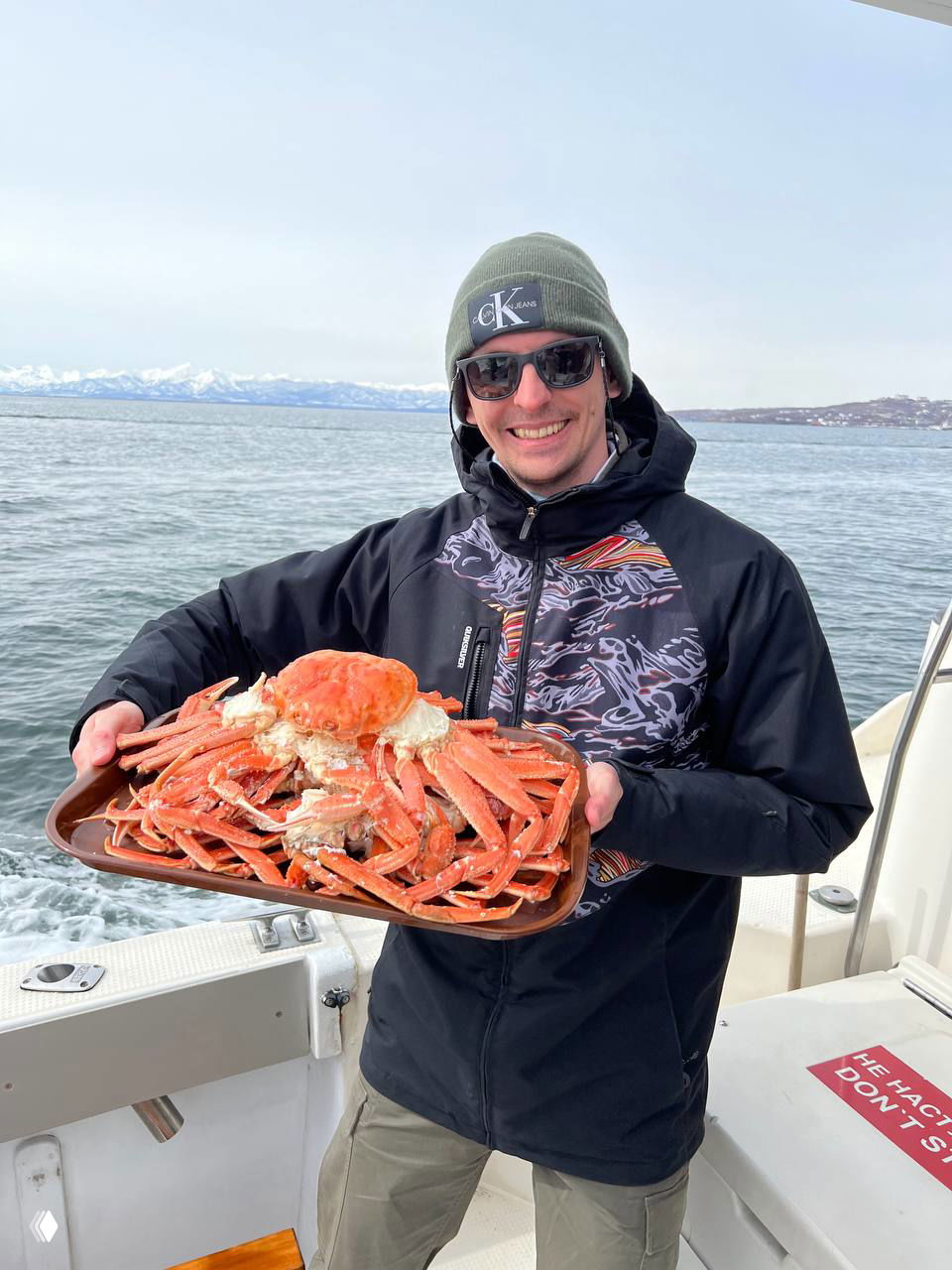 Автор в тёплой куртке держит большую тарелку с камчатским крабом на палубе лодки, улыбка на фоне моря и гор.