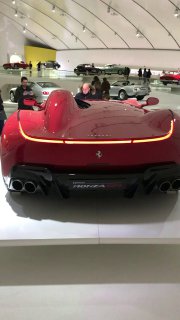 Задняя часть современной модели Ferrari внутри музея, вид на агрессивный диффузор и световую линию заднего фонаря на подиуме.