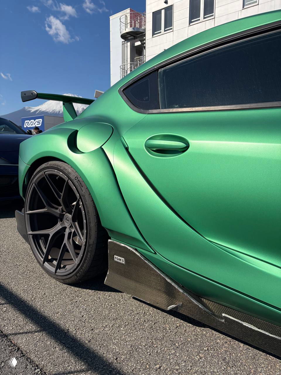 Крупный план боковой части зелёной Supra: расширенное крыло, массивное колесо, карбоновые пороги TOMS и текстура асфальта на пит‑лейне.