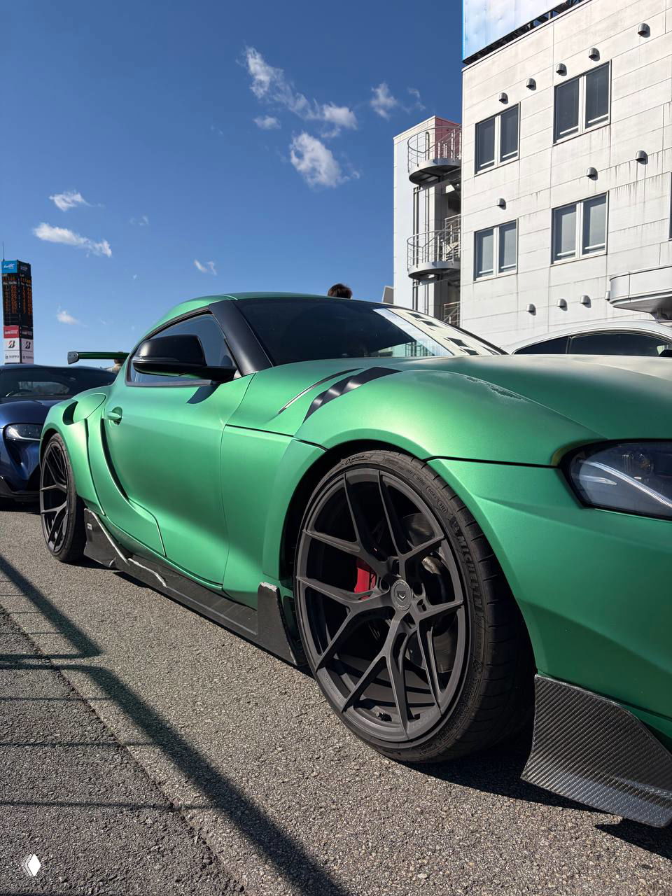 Фронтальный трёхчетвертной вид зелёной Toyota Supra TOMS на парковке у трассы, видно чёткие линии кузова, большие колёса и синее небо.