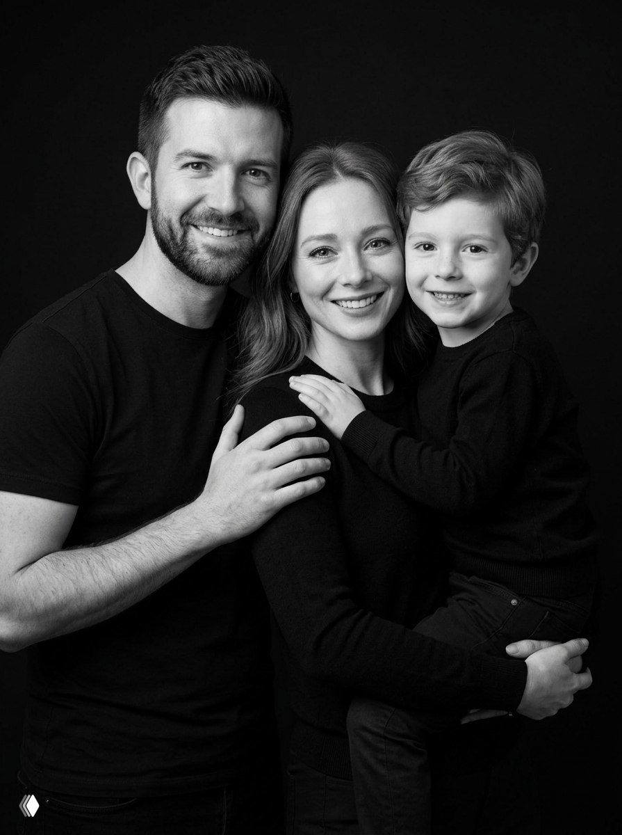 Чёрно-белая студийная фотография семьи: родители и ребёнок в тёмной одежде, плотная групповая композиция, искренние улыбки и мягкие тени на лицах.