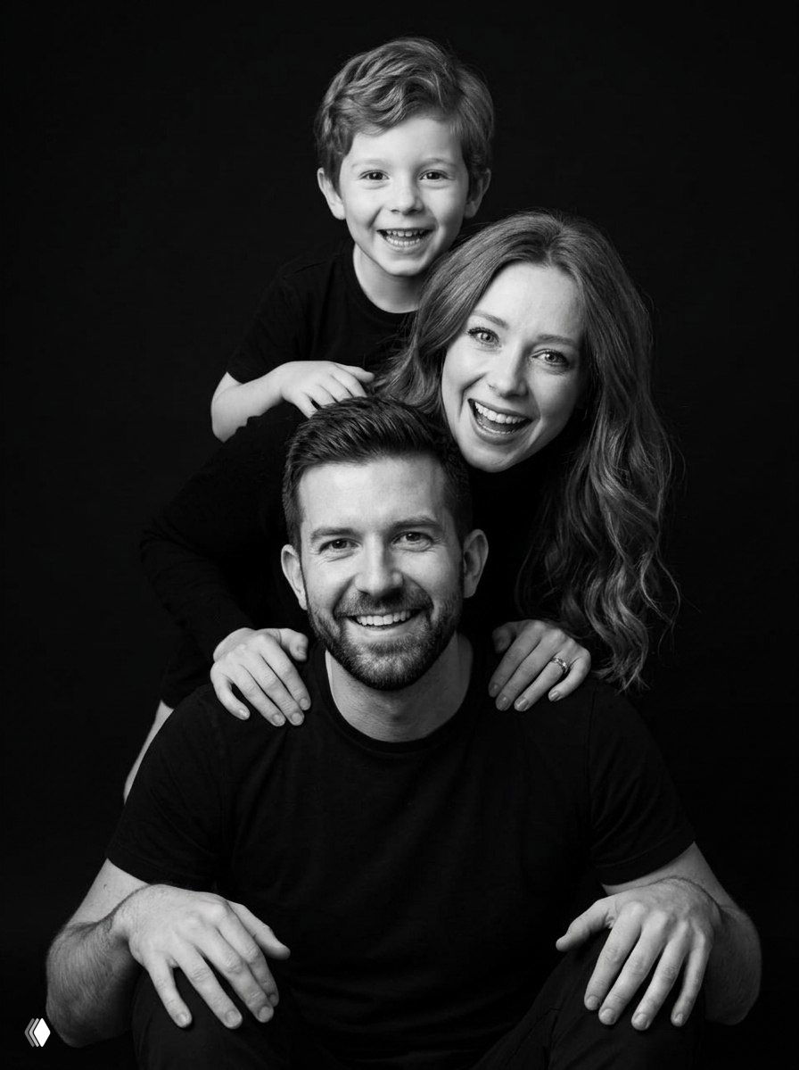 Чёрно-белый студийный портрет семьи на глубоком чёрном фоне: отец, мать и сын близко друг к другу, тёплые улыбки и мягкий направленный свет, крупный план.