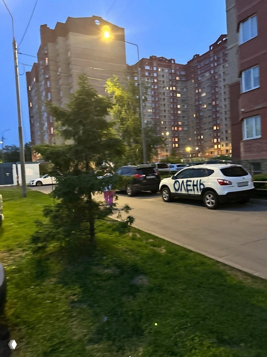 Белая машина во дворе с крупной надписью «ОЛЕНЬ» баллончиком на борту, вечерняя подсветка и многоэтажки на заднем плане — пример вандального промта.