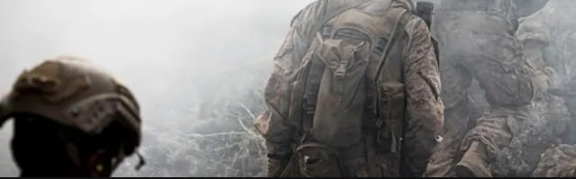 Группа солдат в боевой экипировке идёт через дымку и пыль на фоне разрушенной местности — иллюстрация к новостям о конфликтах.
