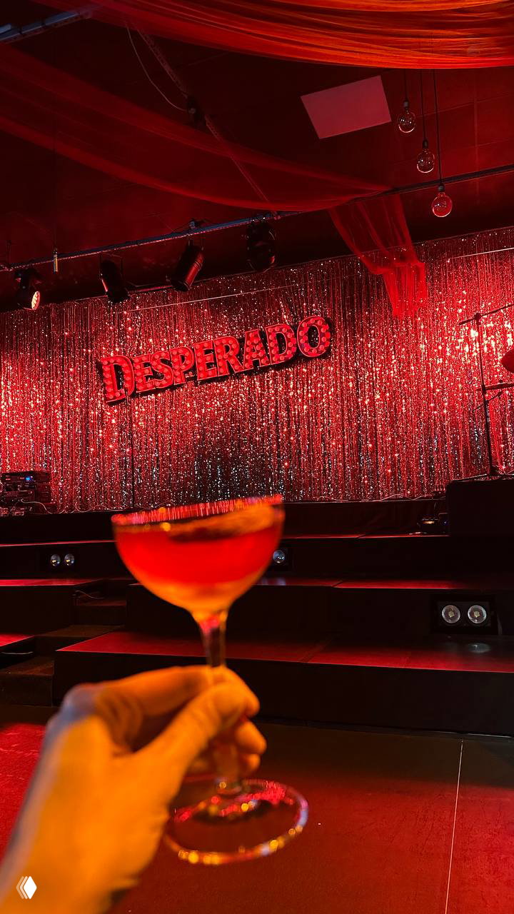 Коктейль вблизи на фоне сцены с красными занавесами и вывеской DESPERADO: атмосферный план зала и сервировки в театральном формате.