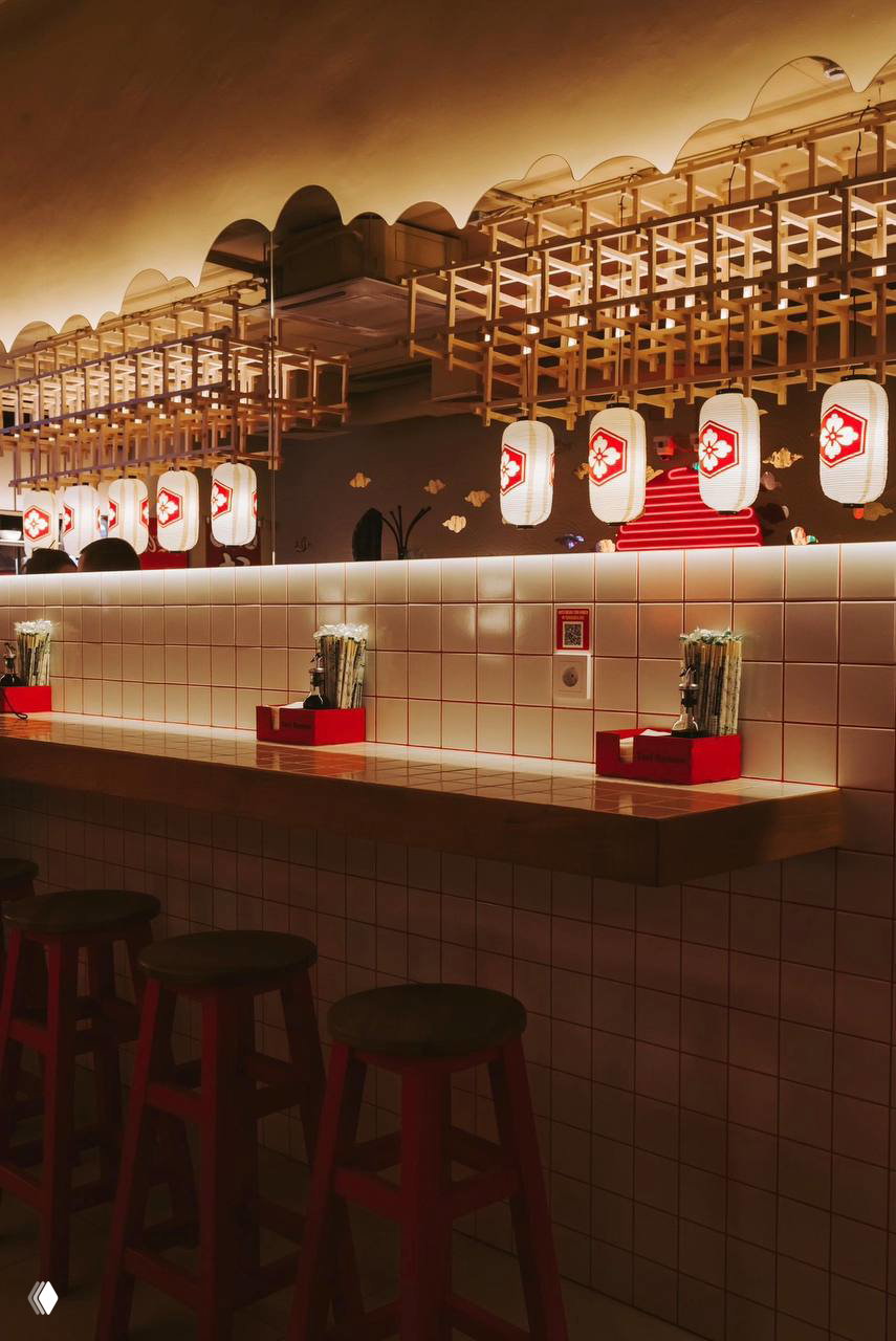 Барная зона Tori Ramen: белая плитка, деревянные подвесные конструкции и бумажные японские фонари над элегантной стойкой с набором палочек и сервировки.