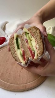 Рука держит разрезанный пополам сэндвич на разделочной доске: видны томатный соус, творожный и лёгкий сыры и свежие овощи в слоях.