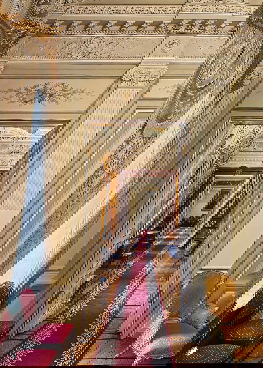 Элегантный коридор и дверные проёмы в здании: длинный красный дорожный ковёр, потолочные росписи, стулья и мягкая подсветка.