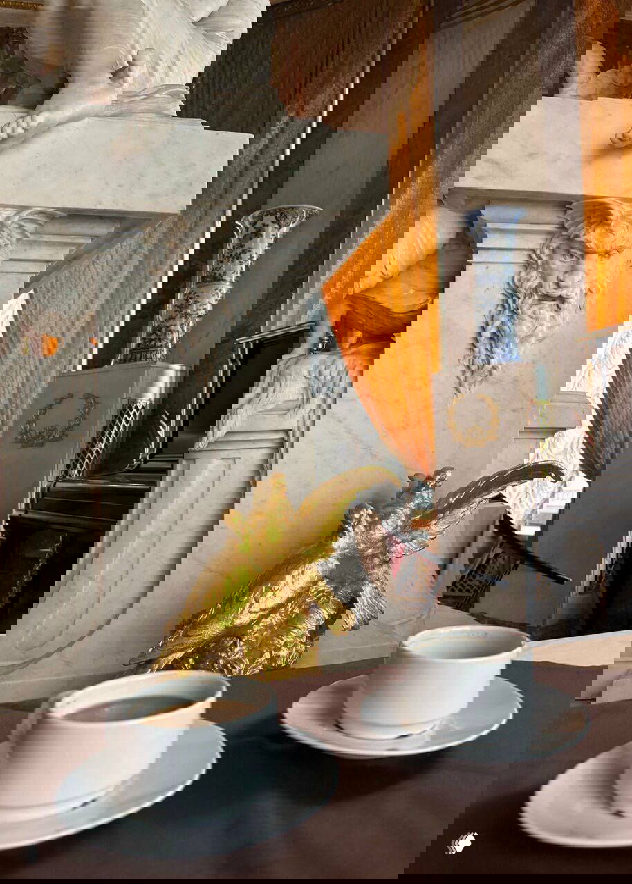 Фрагмент мраморного фонтана со скульптурой, позолоченные детали и чашки кофе на столе у окна с золотыми шторами.