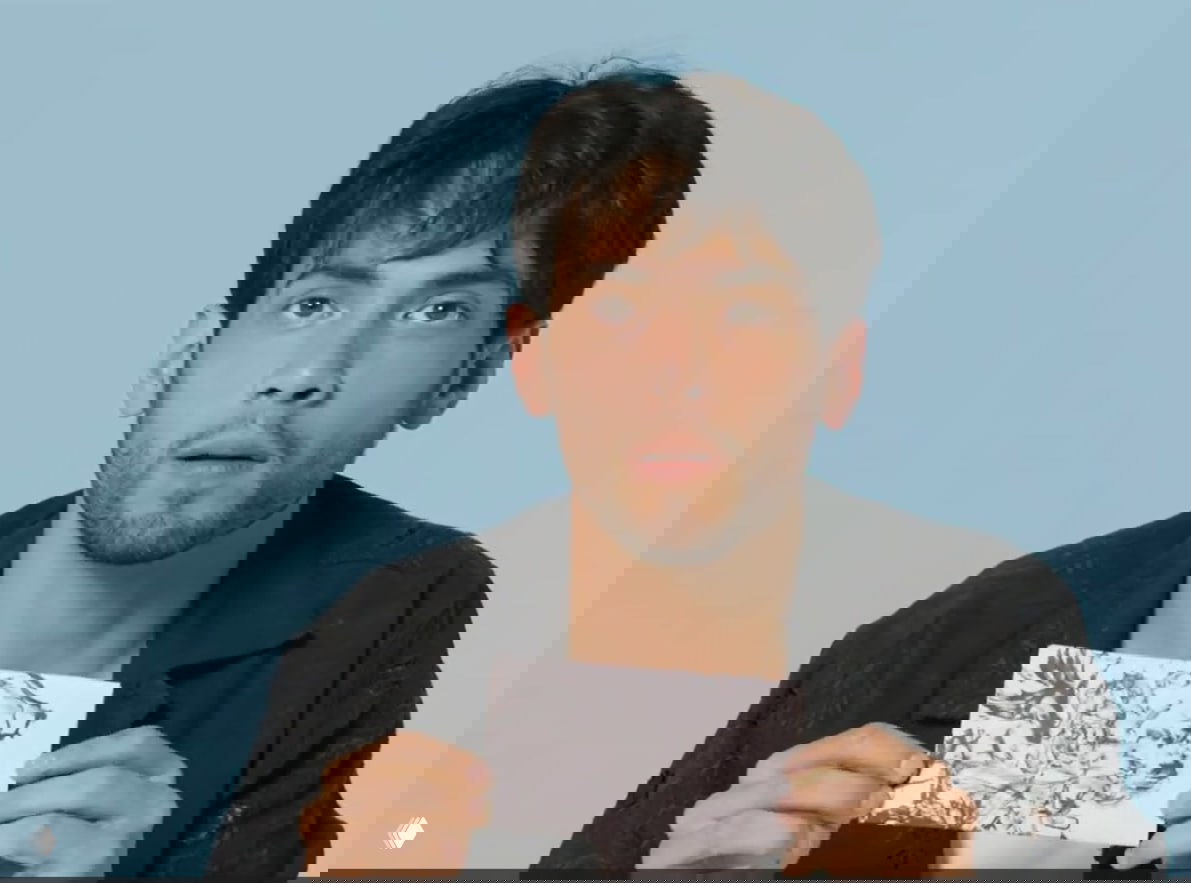 Портретный кадр: мужчина держит черно‑белую зарисовку на бумаге на фоне светло‑голубой стены, смотрит в кадр