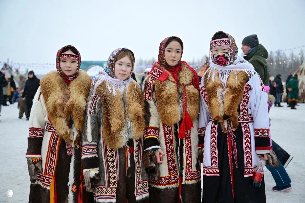 Четыре девушки в традиционных северных шубах и узорчатых платках стоят вместе на снегу перед зрителями во время празднования.