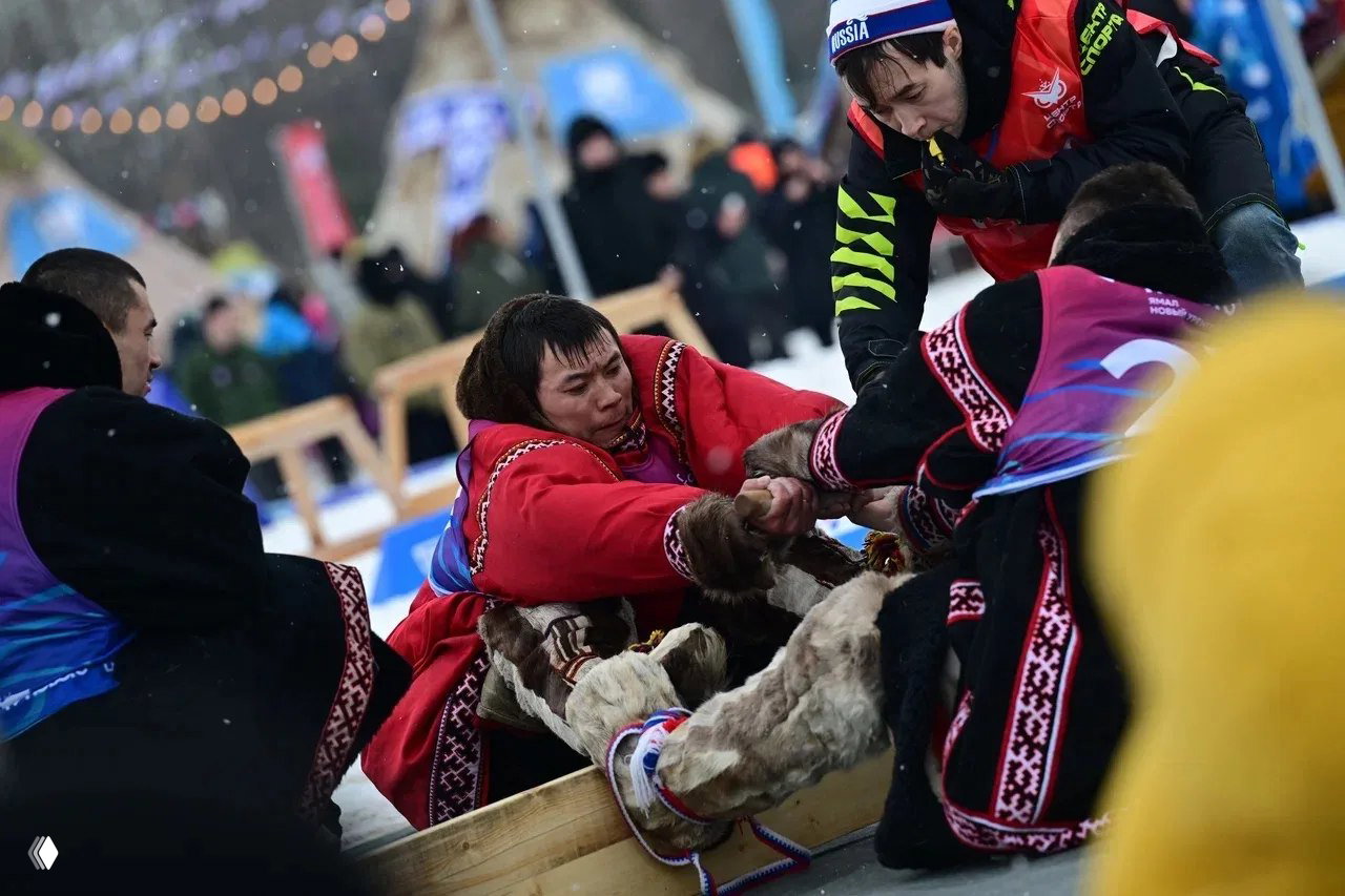 Участники состязания тянут сани в традиционных нарядах на фоне зрителей — напряжённый момент народной игры на льду и снегу.