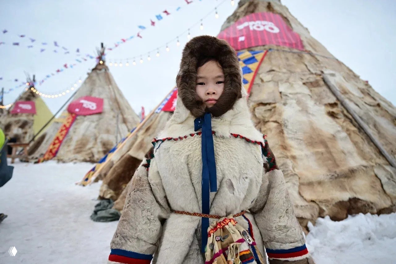 Ребёнок в тёплой традиционной меховой парке крупным планом на фоне чумов и праздничной ярмарки в северной деревне.