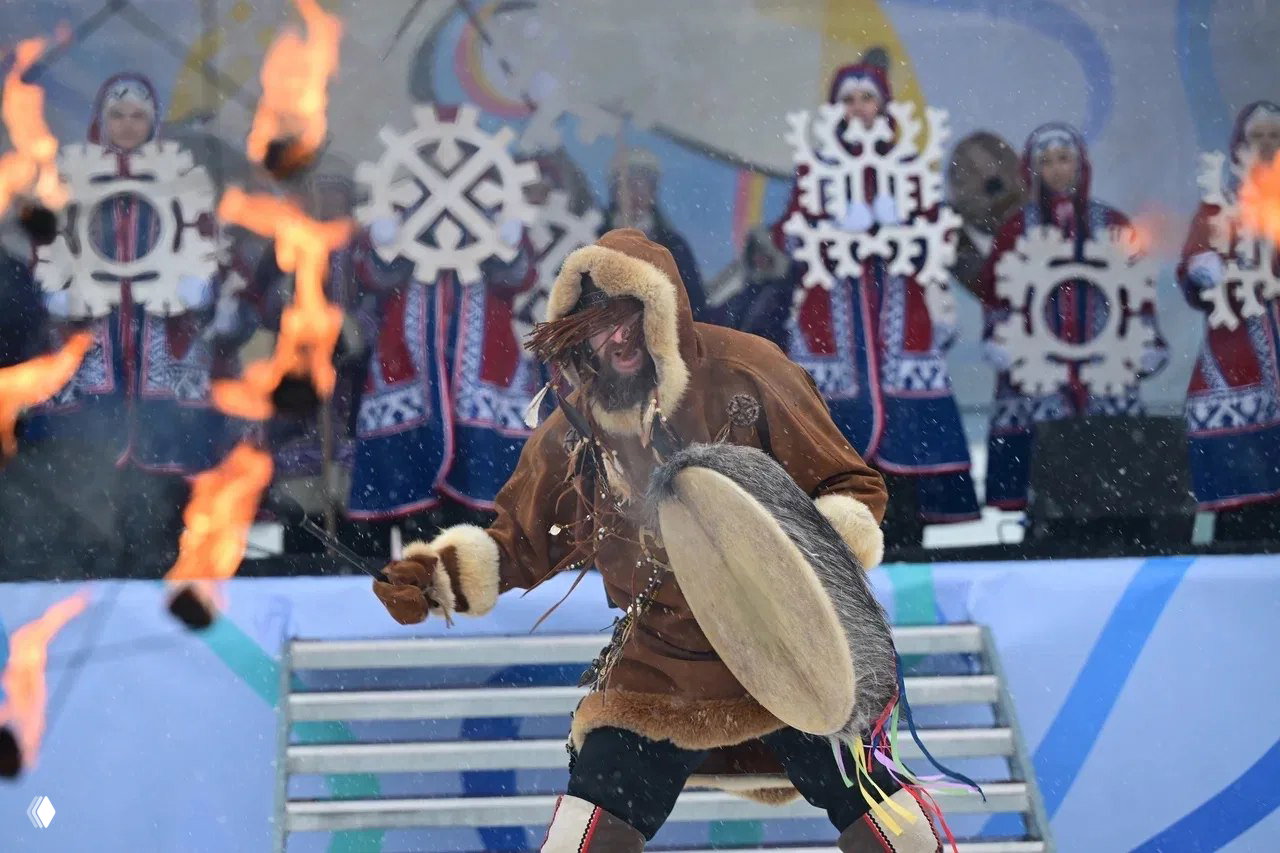 Артист в меховой одежде бьёт в большой бубен на сцене, вокруг — участники хора в национальных костюмах и языки пламени.