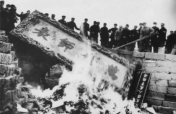 Чёрно‑белая фотография с горящими вывесками и людьми на стене — сцена коллективного уничтожения символов культуры того периода.