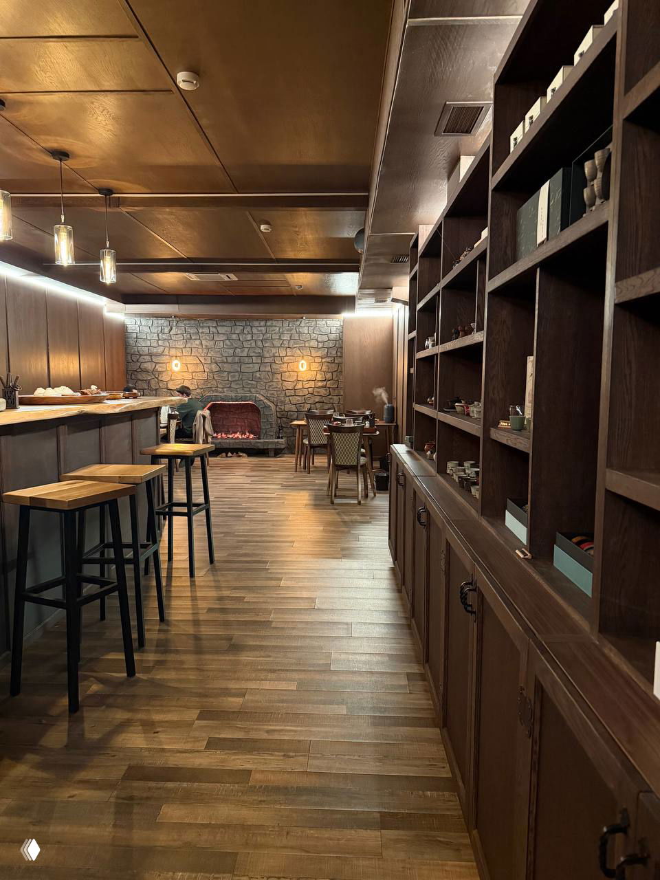 Вид на основной зал с барной стойкой, деревянным полом и стеллажами с чайной посудой; тёплые лампы и каминная ниша на заднем плане.