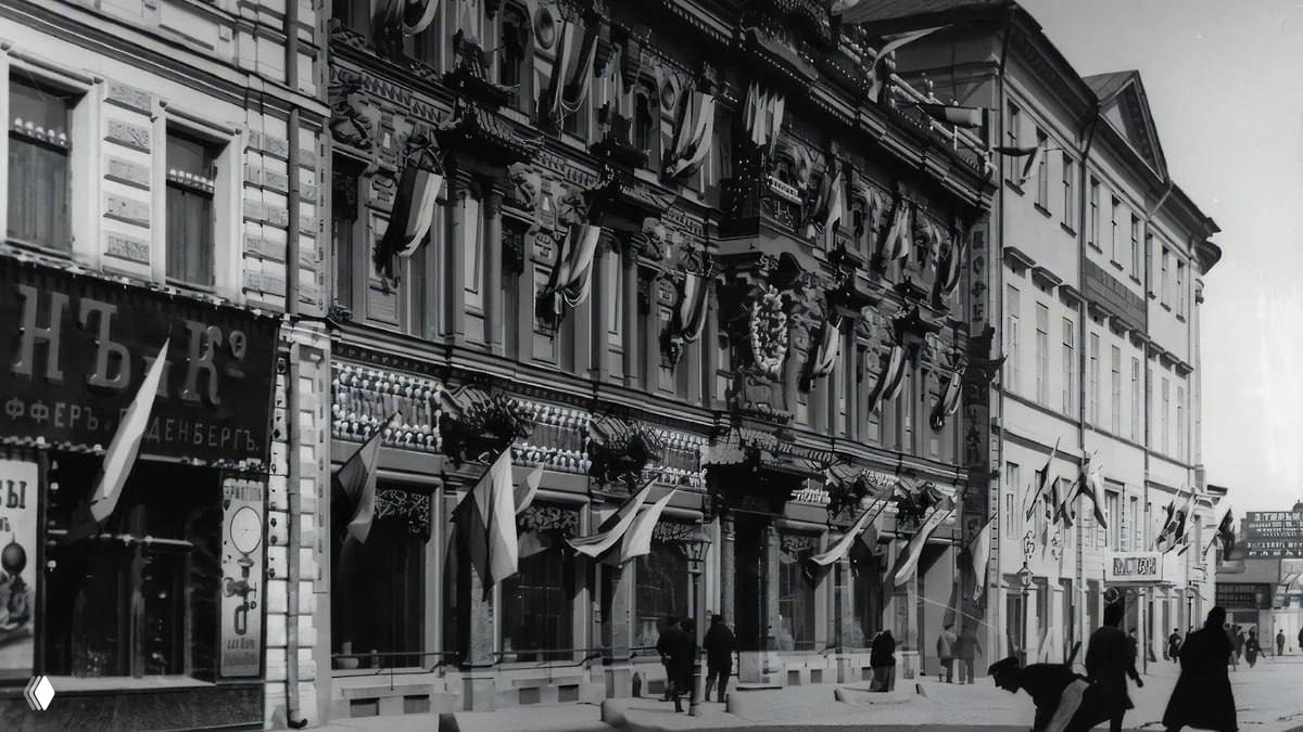 Чёрно‑белая историческая фотография фасада Перлова на Мясницкой с праздничными флагами и прохожими у витрин конца XIX — начала XX века