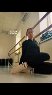 Женщина в спортивной одежде сидит на полу в студии у станка, демонстрирует упражнение для мобильности таза и осанки.