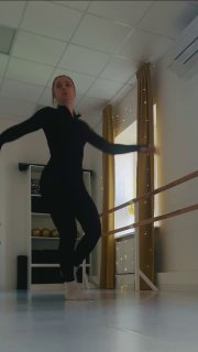 Женщина в тёмном спортивном костюме выполняет прыжок у балетного станка в светлом зале, демонстрируя силу, координацию и работу мышц ног.