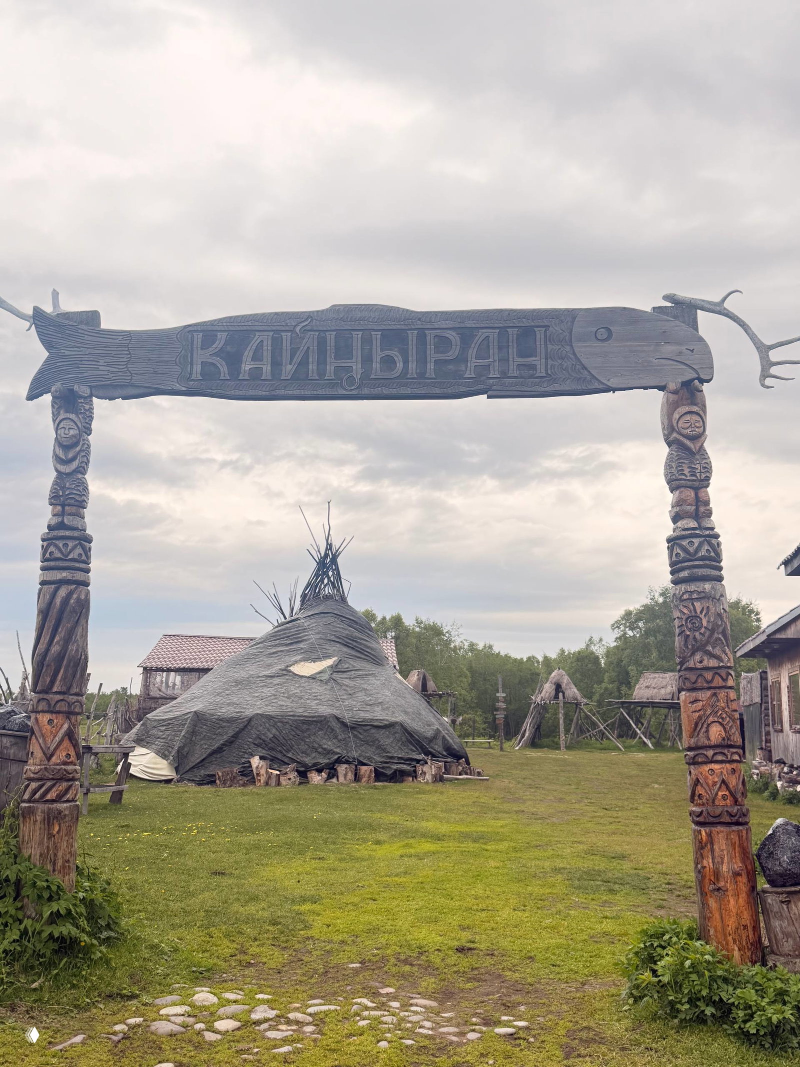 Резные деревянные ворота с надписью «КАЙНЫРАН» на фоне яранги и открытого поля — входная арка этнодеревни и вид на традиционные жилища.