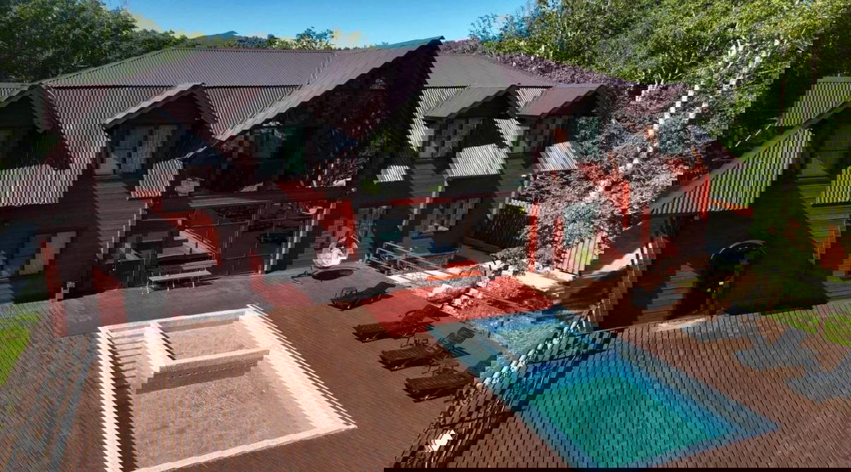 Снаружи деревянный гостевой дом у леса с большой террасой и открытым бассейном — вид на зону отдыха и прозрачную воду.