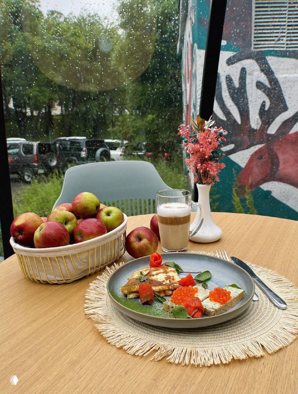 Стол у окна с корзиной яблок, тарелкой закуски с икрой и чашкой кофе на фоне дождливого вида на парковку и зелёные деревья.