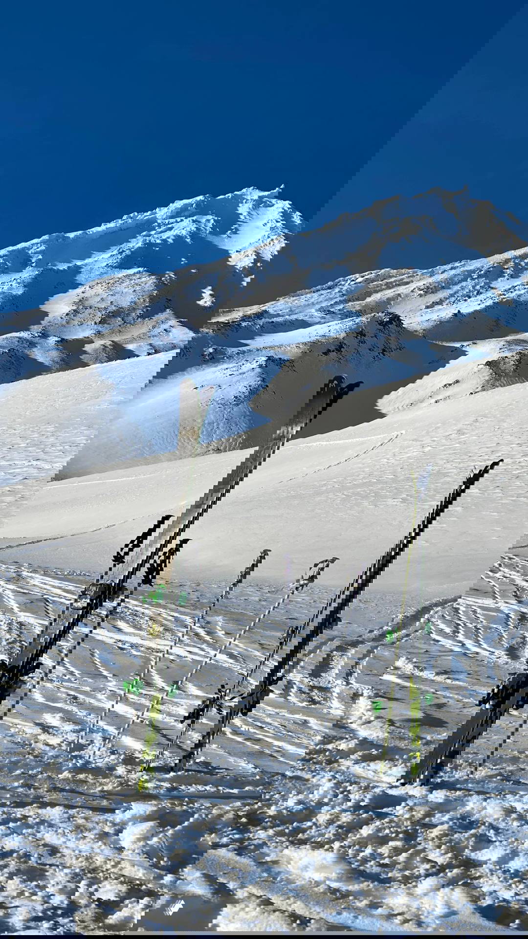 Лыжи и палки, воткнутые в снег на фоне заснеженных гор и яркого синего неба у подножия вулканического гребня на Камчатке.