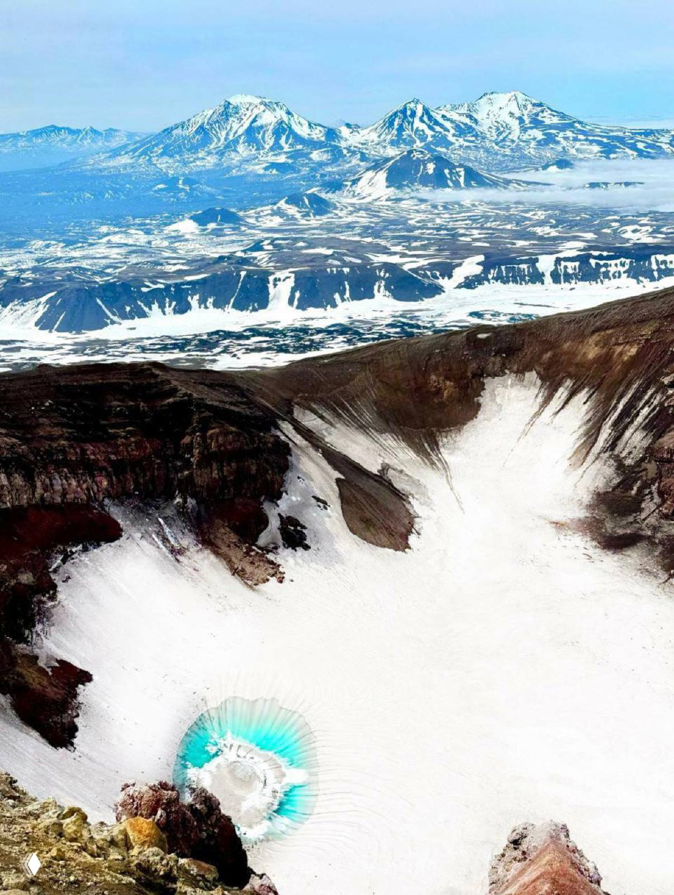 Аэрофото кратера Горелого и окружающих заснеженных вулканических пейзажей Камчатки: маленькое бирюзовое озеро и дальние горы.