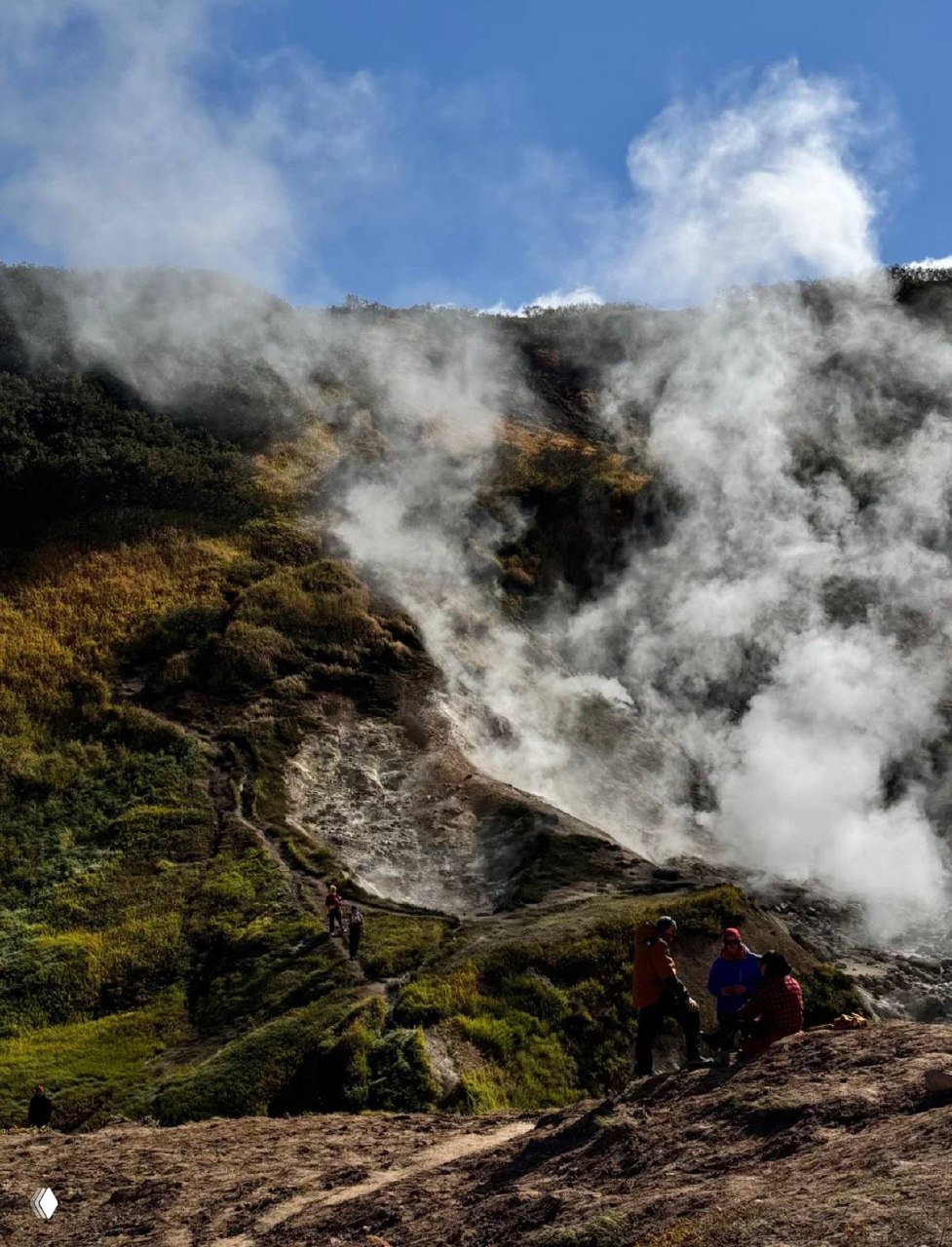 Пар и дым над геотермальной поверхностью Малой Долины гейзеров, тропы и группа туристов на фоне зелёных склонов и голубого неба.