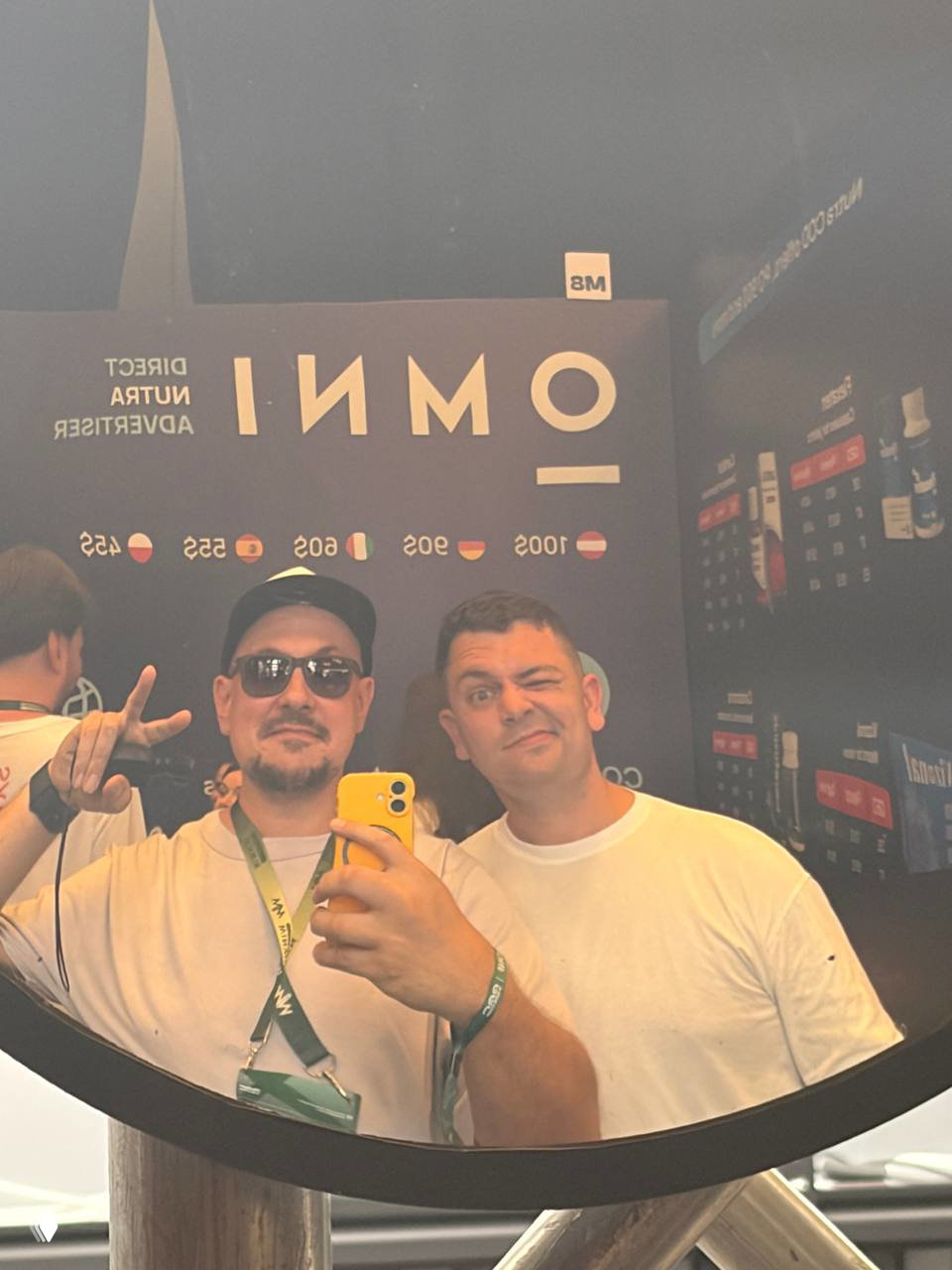 Зеркальное селфи двух мужчин у стенда OMNI на G Gate Conf: один держит жёлтый телефон, оба в бейджах и белых футболках, видно оформление стенда.